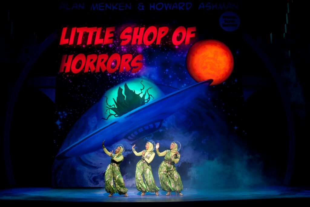 Three women dressed entirely in green sing and dance in front of a picture of a flying saucer with a giant plant inside it. Above it a sign reads "Little Shop of Horrors."