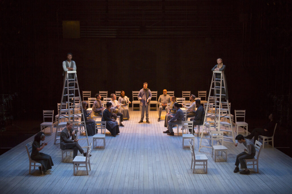 A group of people sit in chairs on a stage. Two stand on tall ladders. One man stands and speaks to the group.
