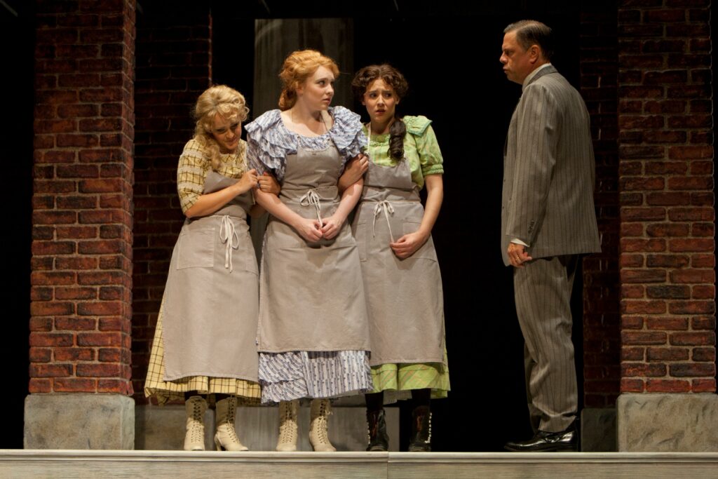 Three scared looking young women stand in front of a brick building and speak to an older man in a suit.