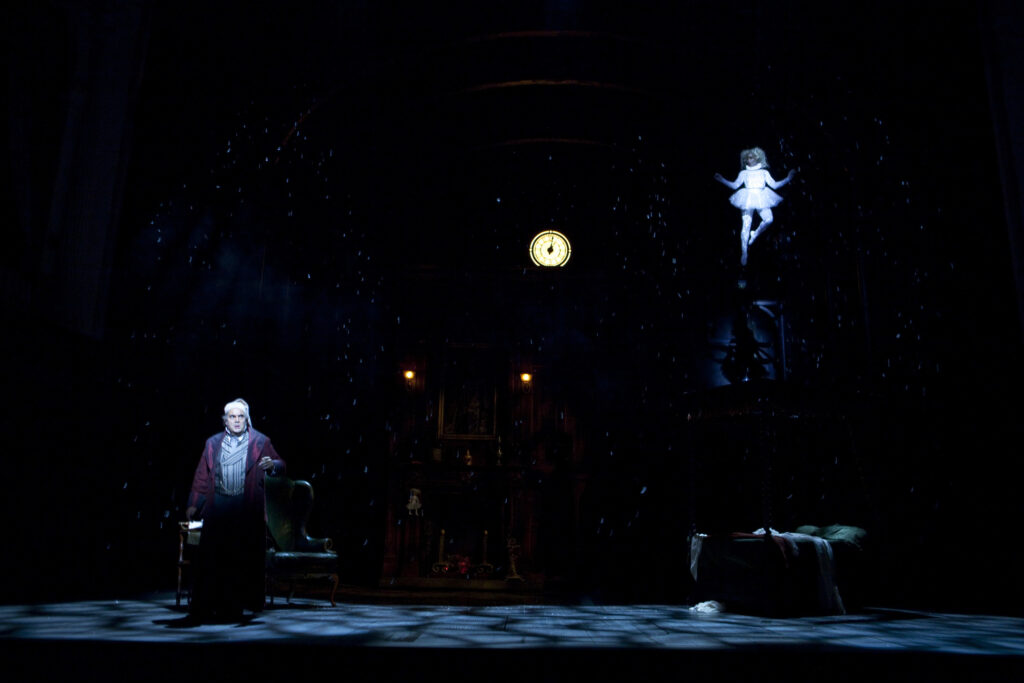 To the left stands Scrooge, an older White man in a white nightcap and nightgown and dark-red silk dressing gown. He faces the camera with a fearful look on his face. Floating high above him in the air to the right is a young Black woman dressed in a sparkling silver tutu, leotard, and tights. Between them is a round yellow clockface which reads 12 o’clock. The lights are darkened, with spotlights on the faces of the two figures. The rest of the set is obscured in darkness.