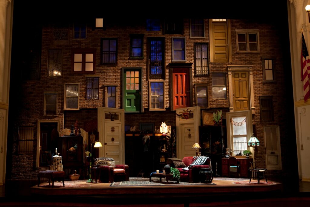 Set design by Tony Cisek is a 2-story exterior brick wall covered in a hodgepodge of window sizes and colorful doors. At the font of the wall sits a living room set that has a red loveseat and armchair, four lamps and various coffee- and side-tables.