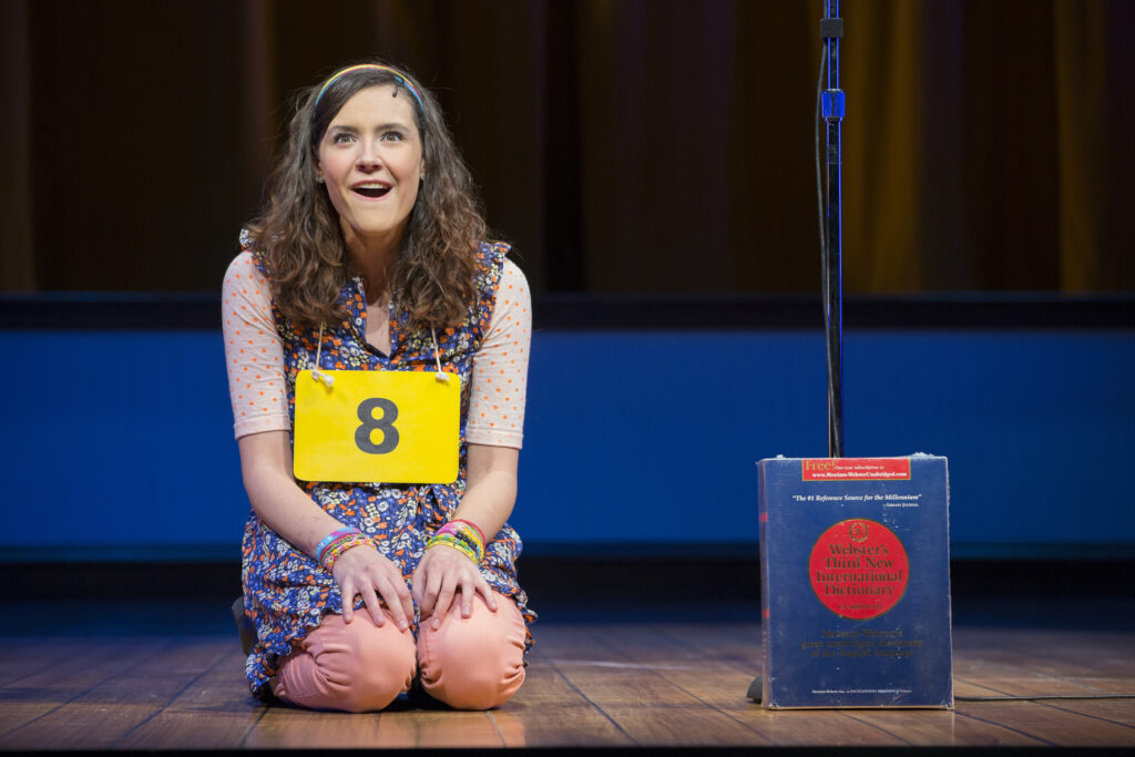 A girl kneels on a stage and smiles. A placard around her neck has the number 8 on it. Next to her a dictionary rests against a microphone stand.