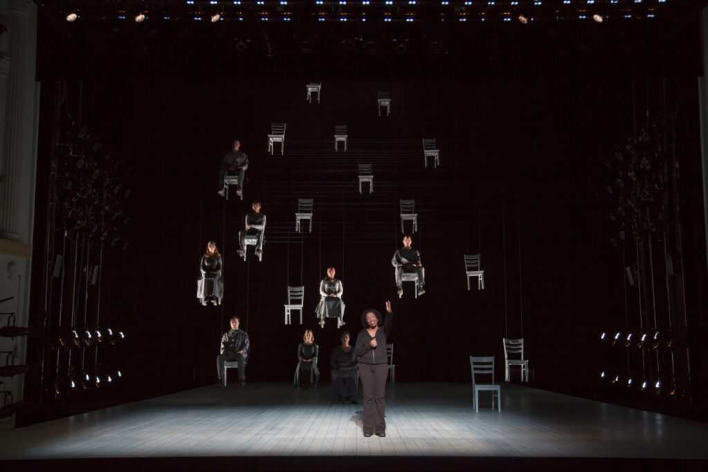 A woman stands on a stage and speaks to the audience. Behind her a number of chairs are suspended in mid-air, half of which have people sitting in them.