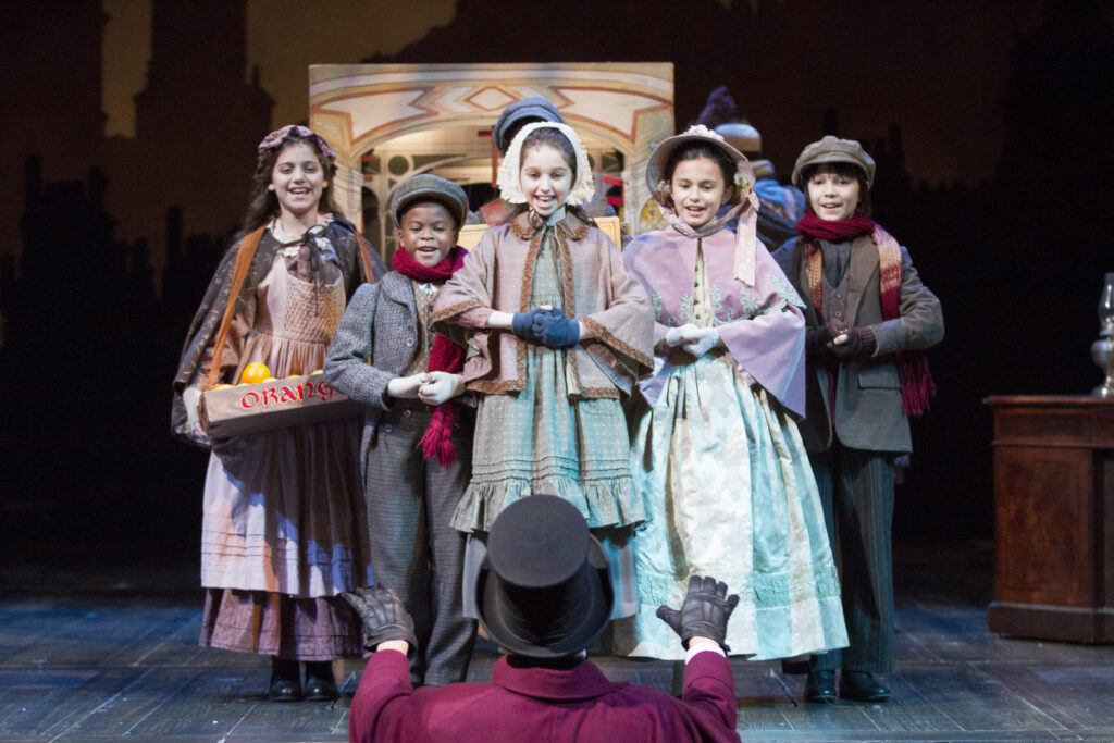 Five children stand and sing under the direction of a man in a purple coat and top hat.
