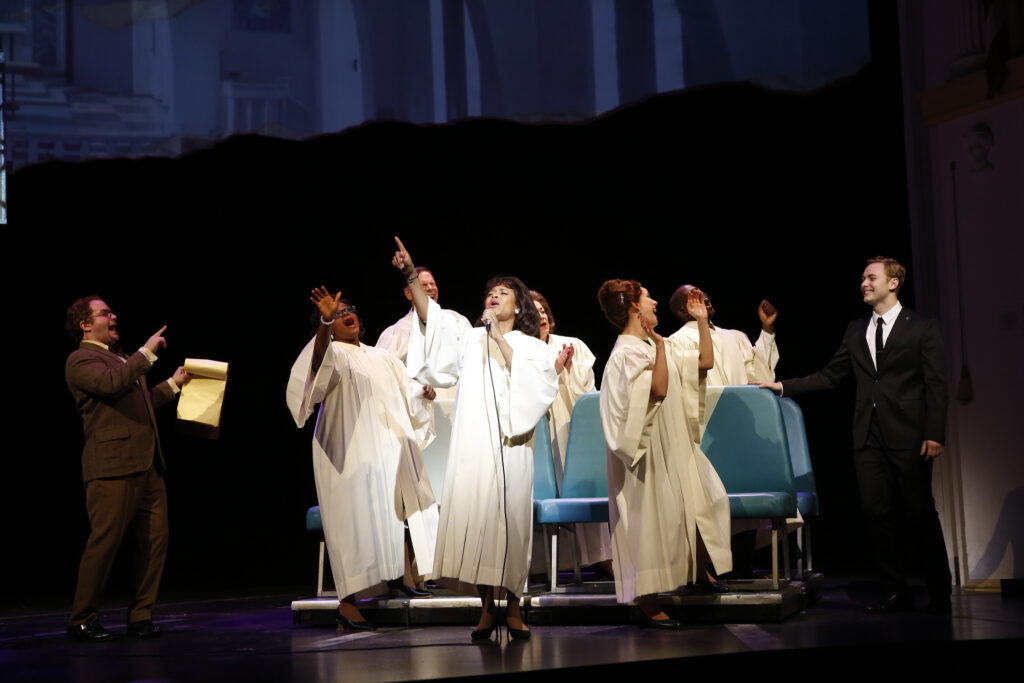 Six people in long white robes sing and dance. To either side men in suits watch and smile.