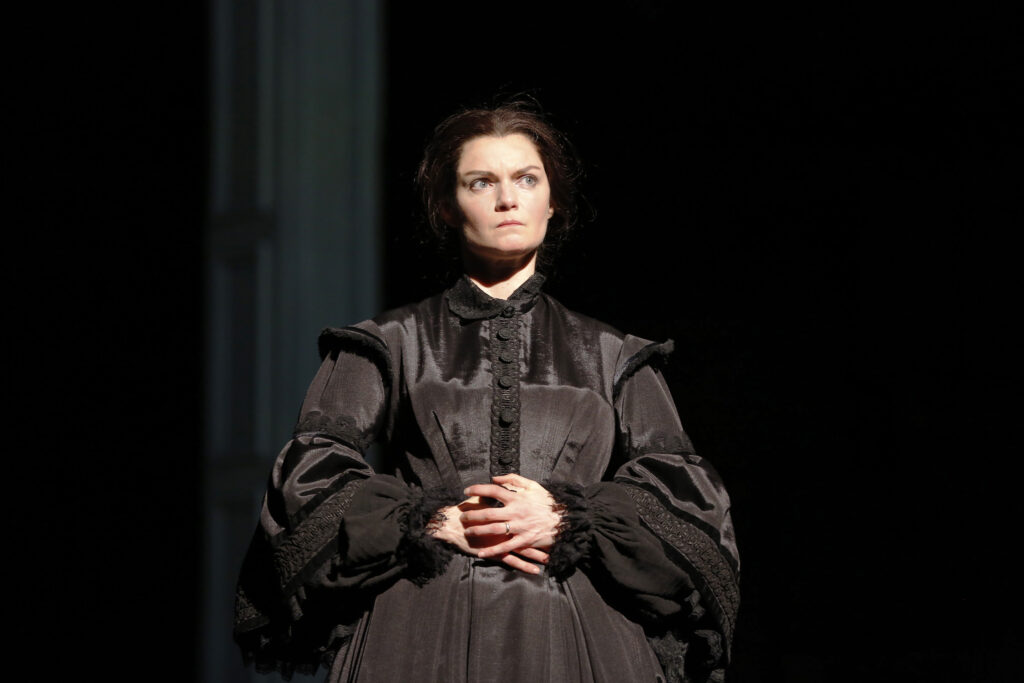 An actress portraying Mary Lincoln stands with her hands around her waist. She is wearing an all-black gown.