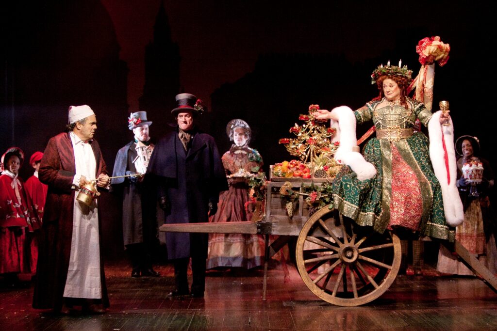 A woman in a green and red embroidered dress sits on a wheelbarrow full of Christmas decorations and holds out her hands while a man in Victorian nightclothes watches her. A group of people in Victorian clothes stand behind them.