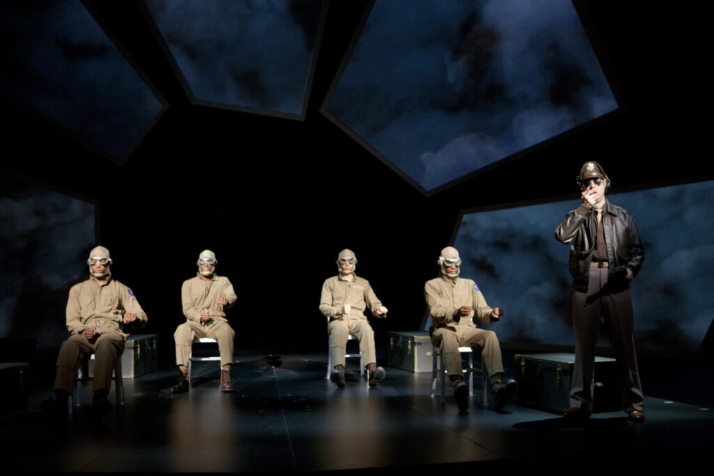 Four African-American men in khaki military uniforms and flight helmets sit and mime piloting a plane, while a white man in an aviator jacket and dark glasses mimes speaking into a radio.
