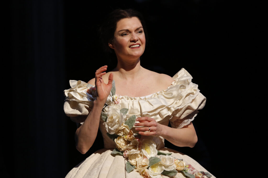 An actress portraying Mary Lincoln waves hesitantly. She is wearing a ruffled cream ballgown with cloth flowers across the chest and waist.
