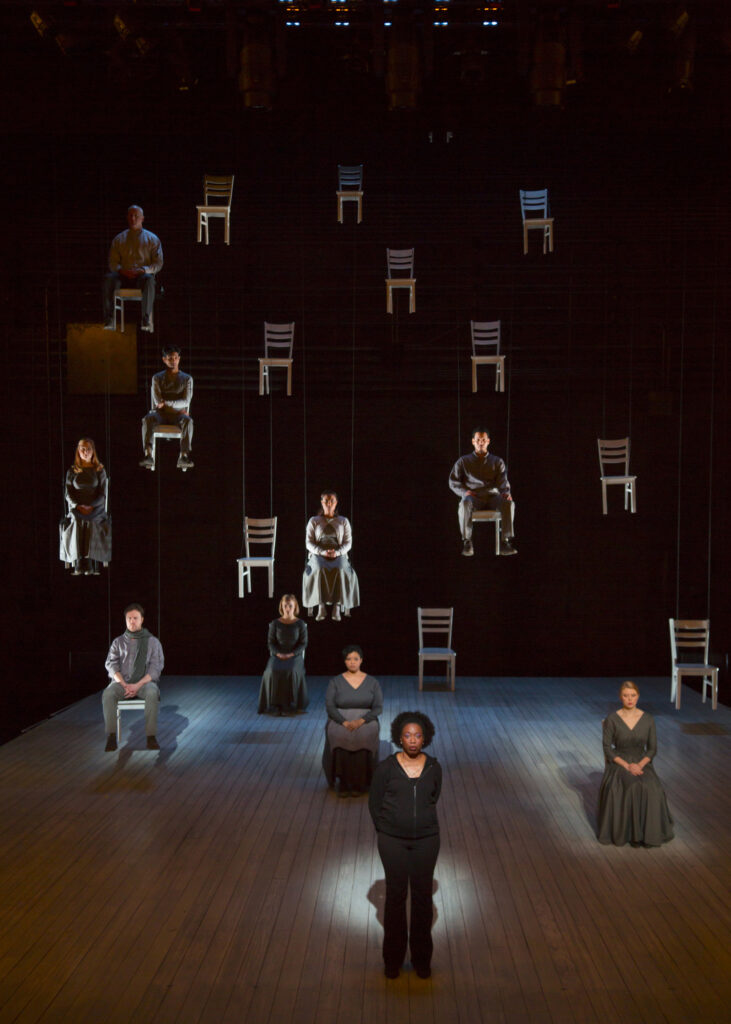 A woman stands on a stage. Several people behind her sit in chairs, and several more sit in chairs suspended by wires in mid-air.