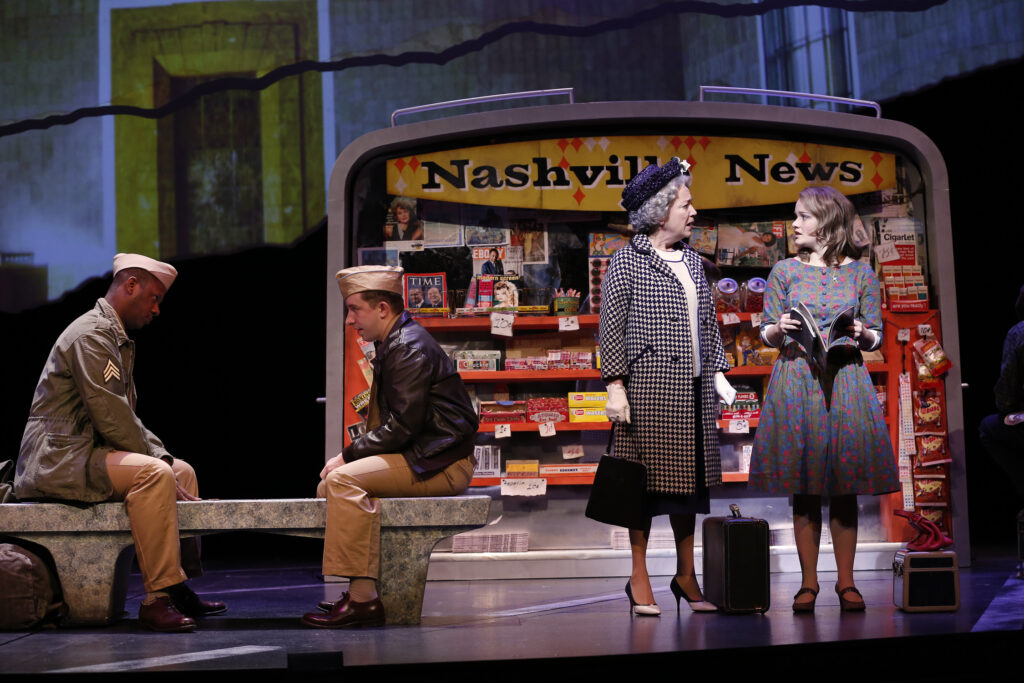Two men in WWII era military uniforms sit on a bench and talk to one another. Next to them a young woman talks to and older woman, both with travelling bags next to them. Behind them is news stand that says "Nashville News" on its sign.