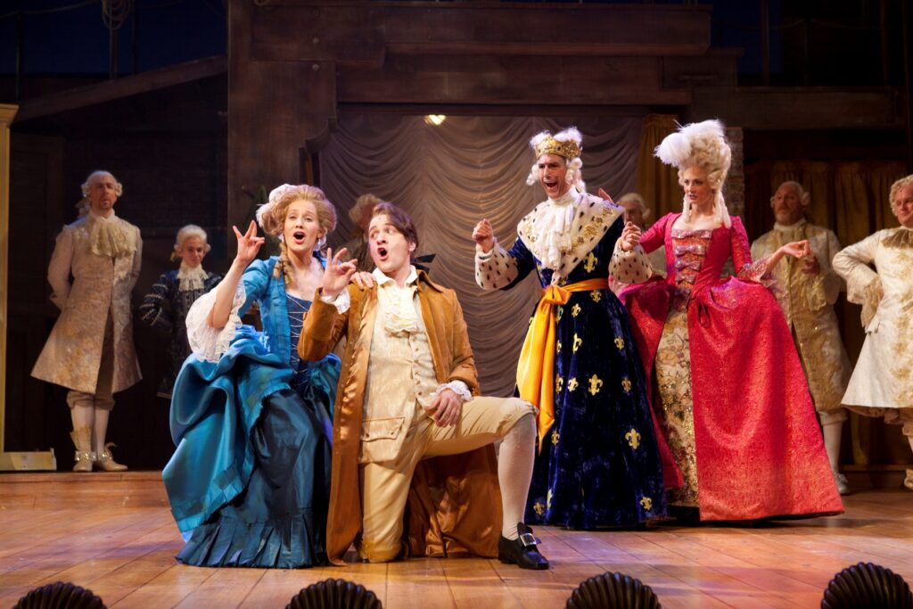 A man and woman in gaudy 18th century clothes kneel on a stage and press their ring fingers and thumbs together with their right hands. Behind them a man in a long blue robe decorated with fleur-de-lis with a crown on his head and a woman in a broad red hoop dress watch them.