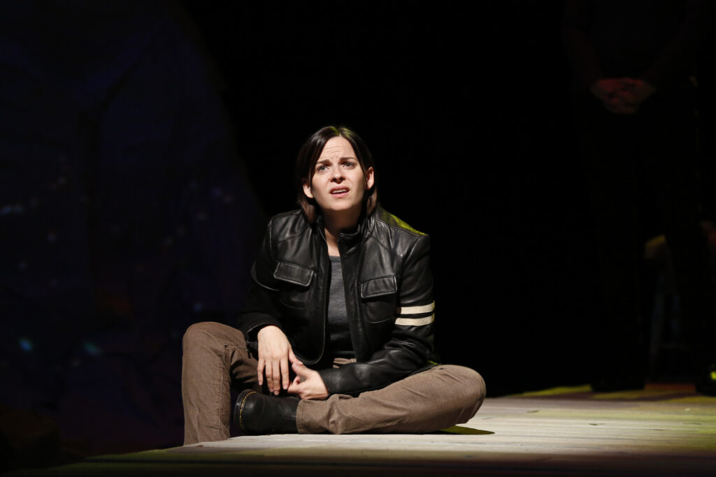 A woman in a leather jacket sits on a wooden floor looking perplexed.