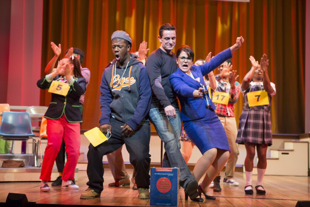 A group of people all dance together on a stage. Most have placards around their necks with numbers on them. In the foreground a dictionary rests on the ground.