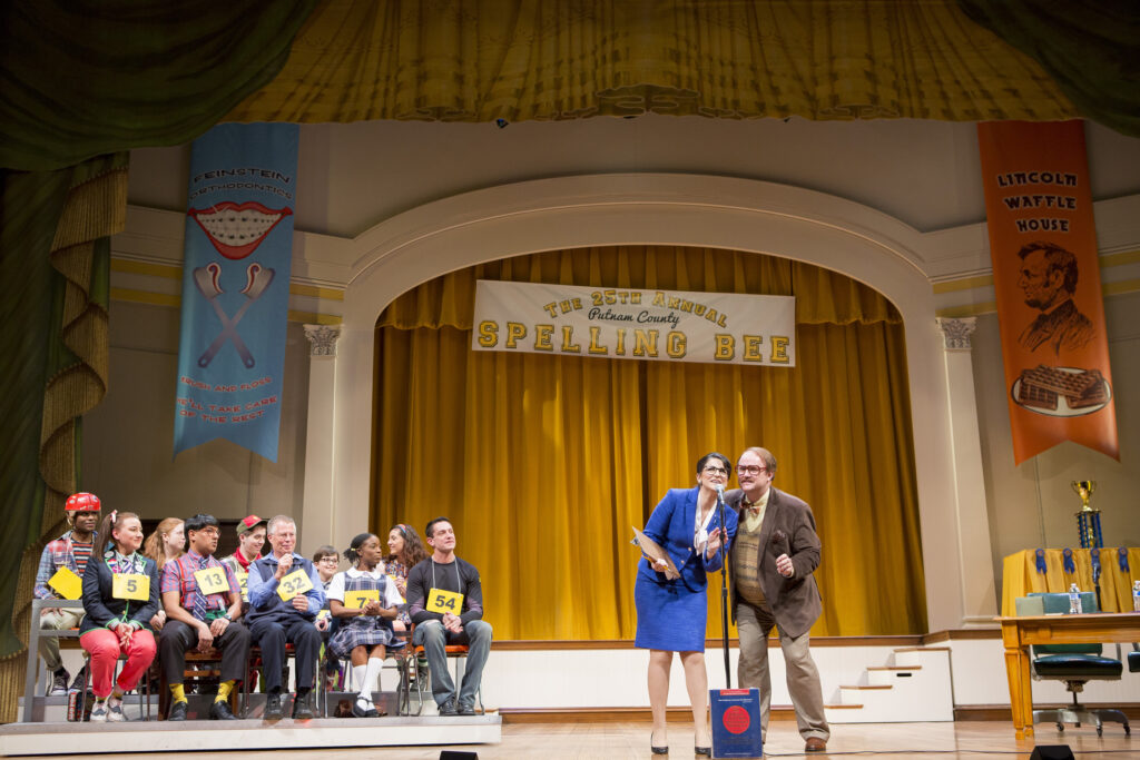 A man in a jacket and bowtie and a woman in a blue blazer speak into a microphone on a stage. Ten actors dressed as children sit off to a side, each with a placard with a number on it around their neck. A banner behind them says "The 25th Annual Putnam County Spelling Bee."