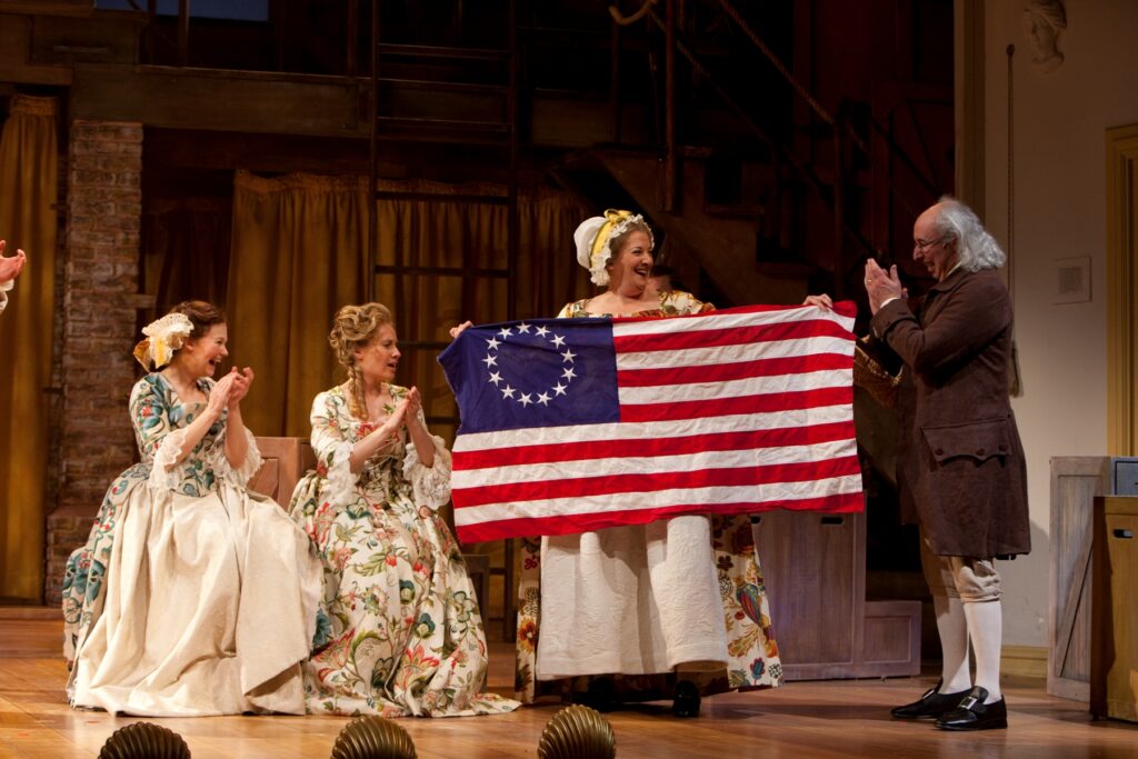 A woman in a white dress with floral patterns holds up an American flag with thirteen stars. Two similarly dressed women sit next to her and applaud. An actor portraying Benjamin Franklin stands and claps as well.