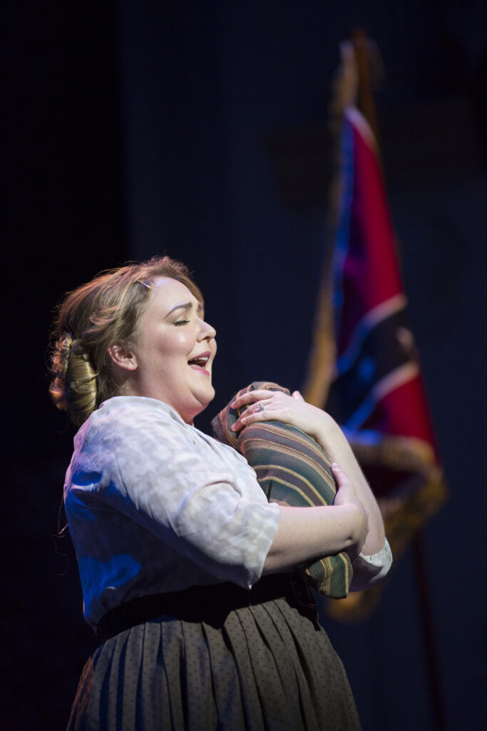 A woman in 19th century clothing holds a swaddled infant and sings. Behind her is a confederate flag.