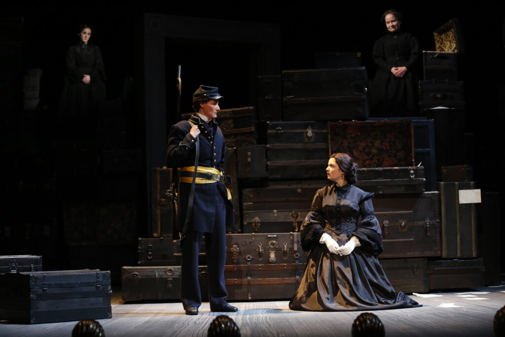 An actress portraying Mary Lincoln, dressed in a black gown and white gloves, kneels on stage. A Union solider in a blue uniform and yellow sash, with a gun behind his shoulder, looks at her. Behind them are two women wearing black.