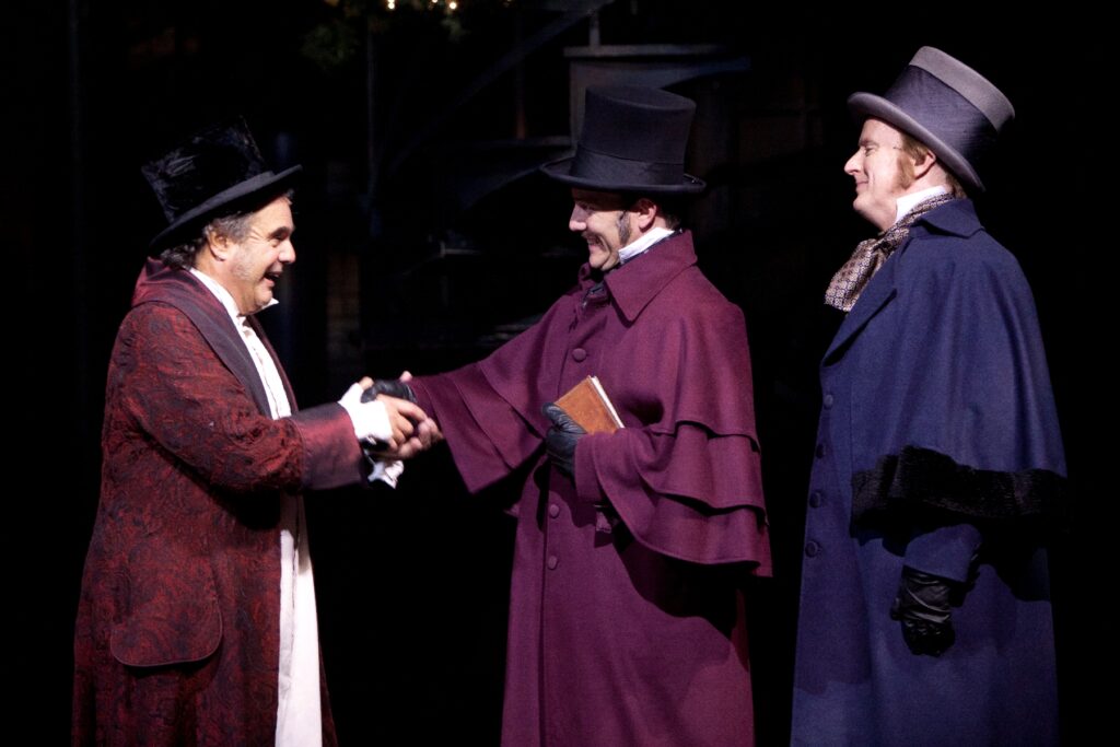 A man in Victorian nightclothes and top hat shakes hands with another man while a third looks on.