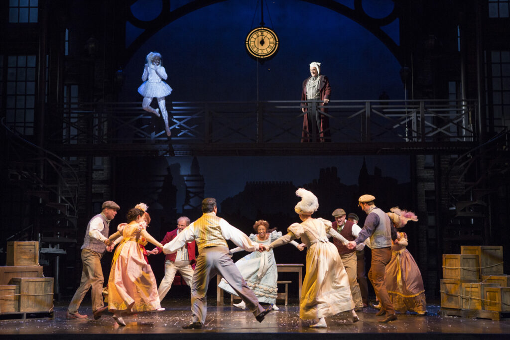 A group of people hold hands and dance in a circle. Above them a woman in white hovers in the air, while next to her a man in his nightclothes stands on a catwalk and watches the dancers.