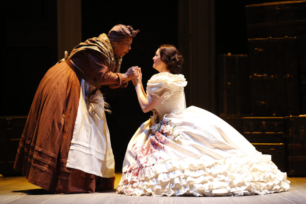 An actress portraying Mary Lincoln kneels on the floor with her hands clutched in the hands of an older African American woman who leans down to her. The older woman is dressed in a simple dress, shawl and headwrap. Mary is dressed in a ruffled cream ballgown with cloth flowers across the chest and waist. There is blood splatter at the bottom of her gown.