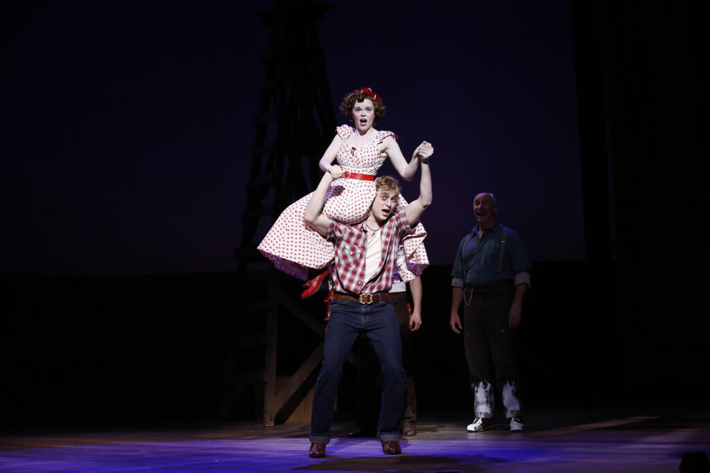 A man in jeans and a plaid shirt holds a woman in a polka dot dress on his shoulder. Behind them a man watches them and smiles.