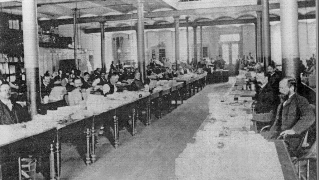 Black and white photograph of a large number of men working at rows of long tables in a spacious office. The tables are full of sheets of paper.