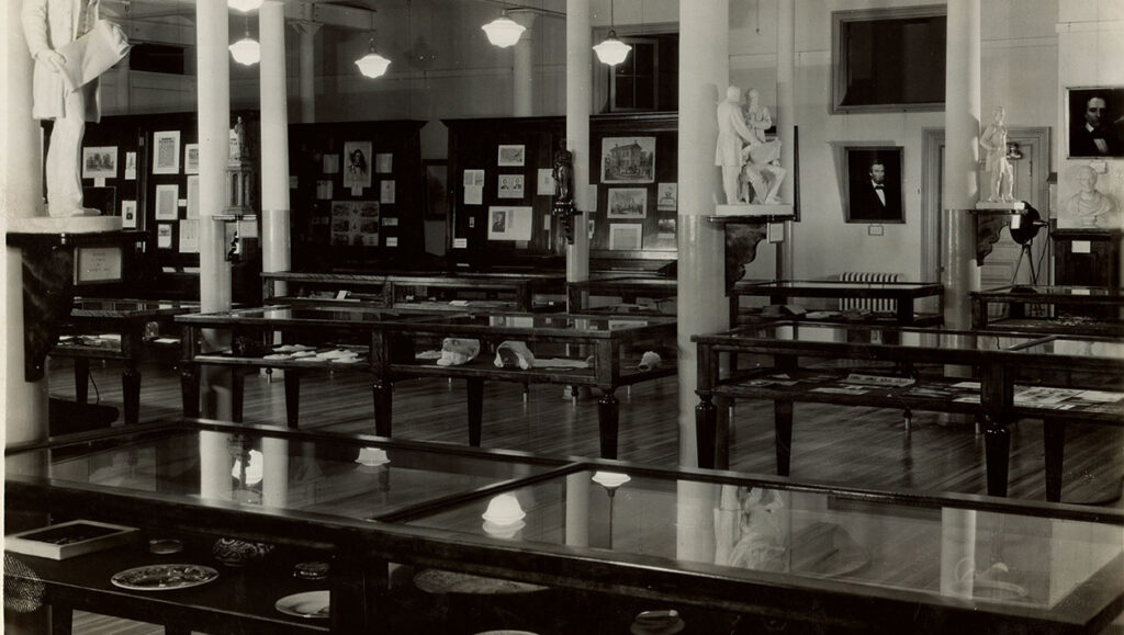 Black and white photograph of a large room filled with display cases and exhibits.