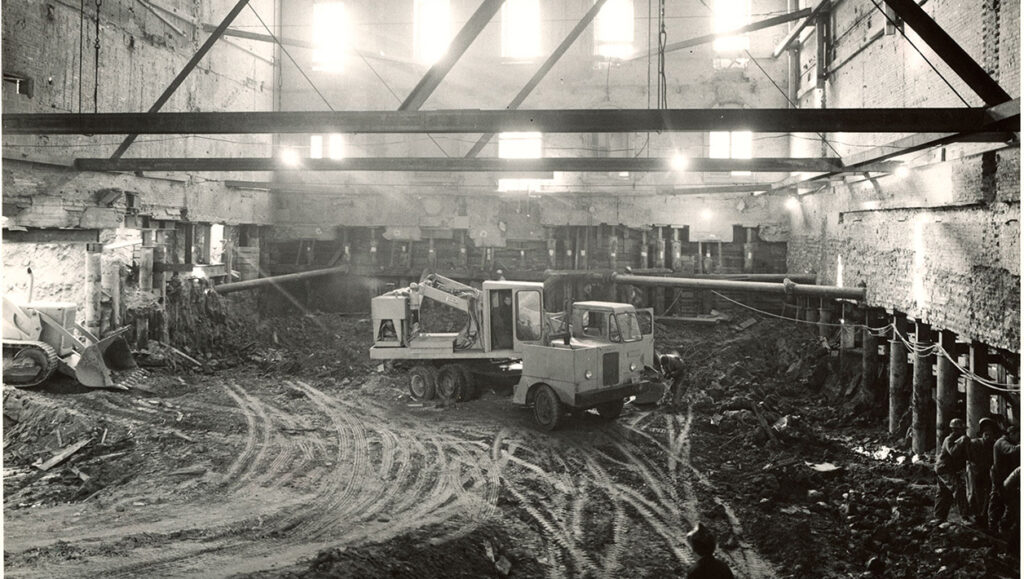 Black and white photograph of the interior of a large building with the floor removed and heavy work machinery operating in the soil underneath.