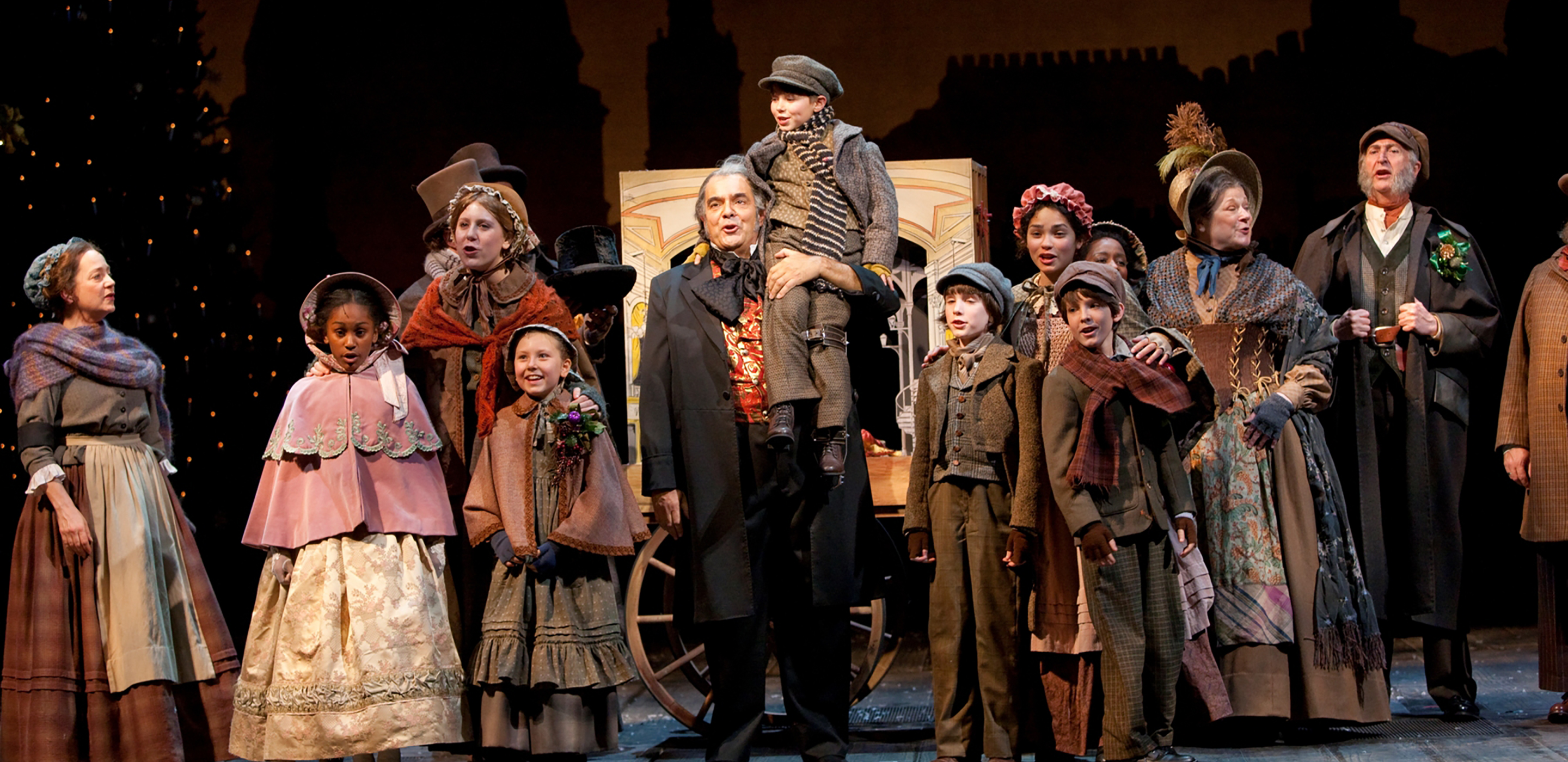Seven children and five adults in Victorian costume surround the actor playing Scrooge on set at Ford’s Theatre. A young boy (playing Tiny Tim) sits on Scrooge’s shoulders.