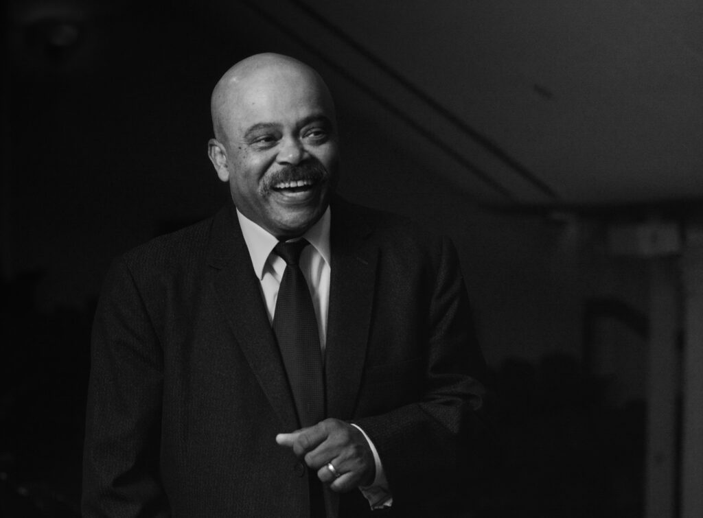 A black and white portrait of Jim Bono. He is an older man who is bald and has a full mustache. He is dressed in a black suit and tie. He is mid-laugh.