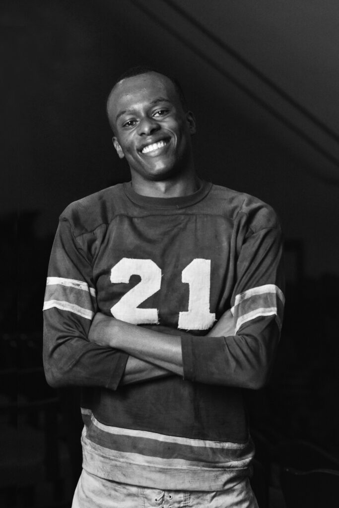 A black and white portrait of Cory Maxson. He is a young man in a dirty football uniform from the 1950s. He has his arms crossed in front of him and smiles broadly for the camera.