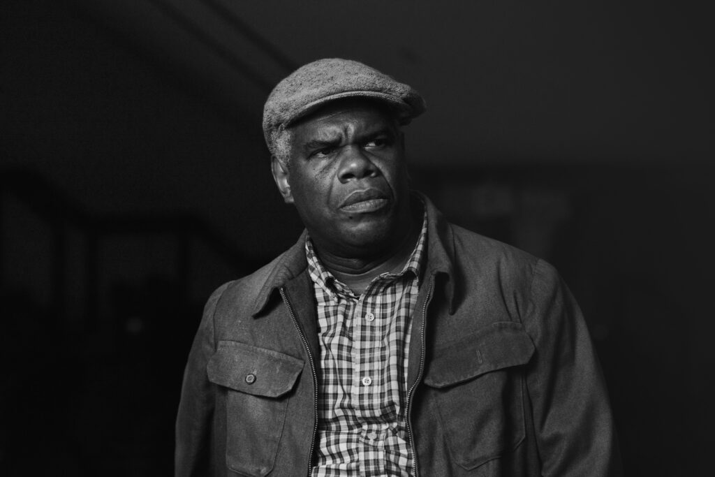 A black and white portrait of Troy Maxson. He is an older man in a slightly battered jacket, cap and checkered work shirt. He grimaces as he looks off into the distance.