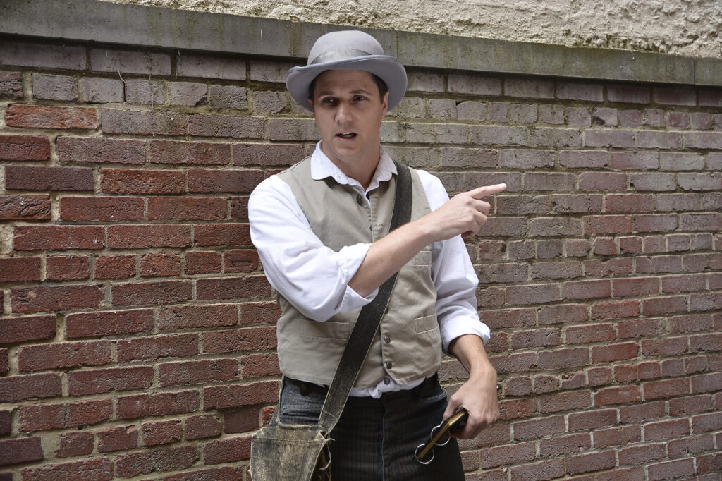 An actor stands dressed as an 1860s detective, holding a notepad. Behind him on a brick wall.