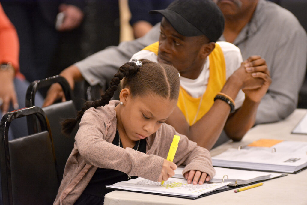Young actress Janiyah Lucas sits at a table with castmates, wearing her hair in pigtails. She highlights her lines in her script during first rehearsal.