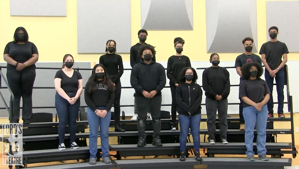 A group of masked teenagers stand on various levels of a set of risers and give a speech.