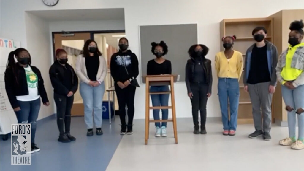 A group of eight masked students stand in a semicircle in a class room. A student behind a desk gives a speech.