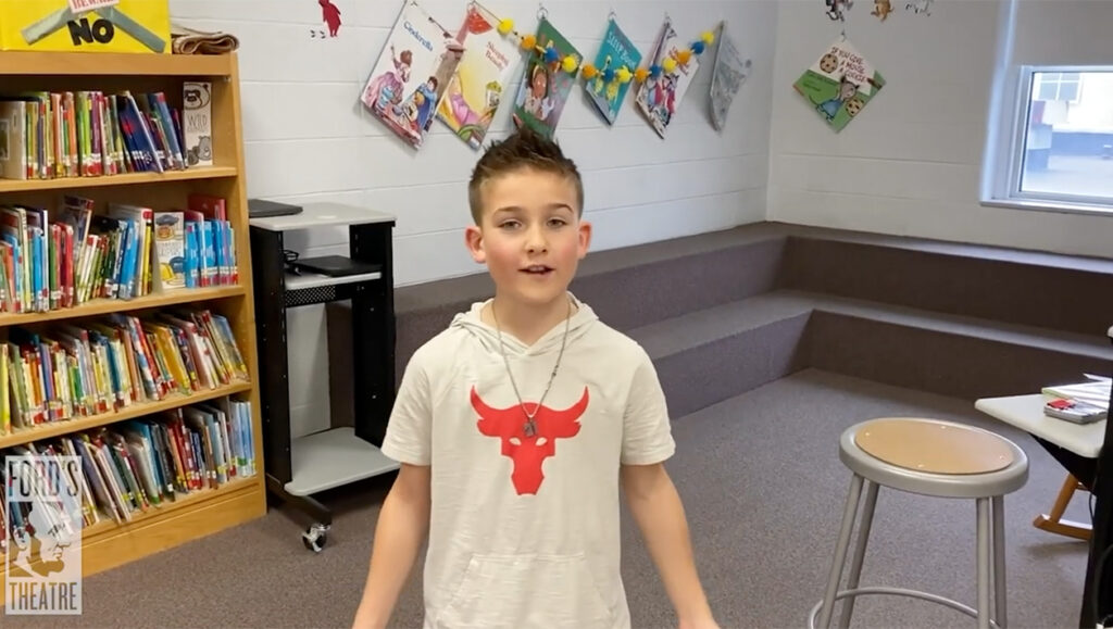 A young boy wearing a t-shirt with a bull emblem stands in a classroom and speaks.