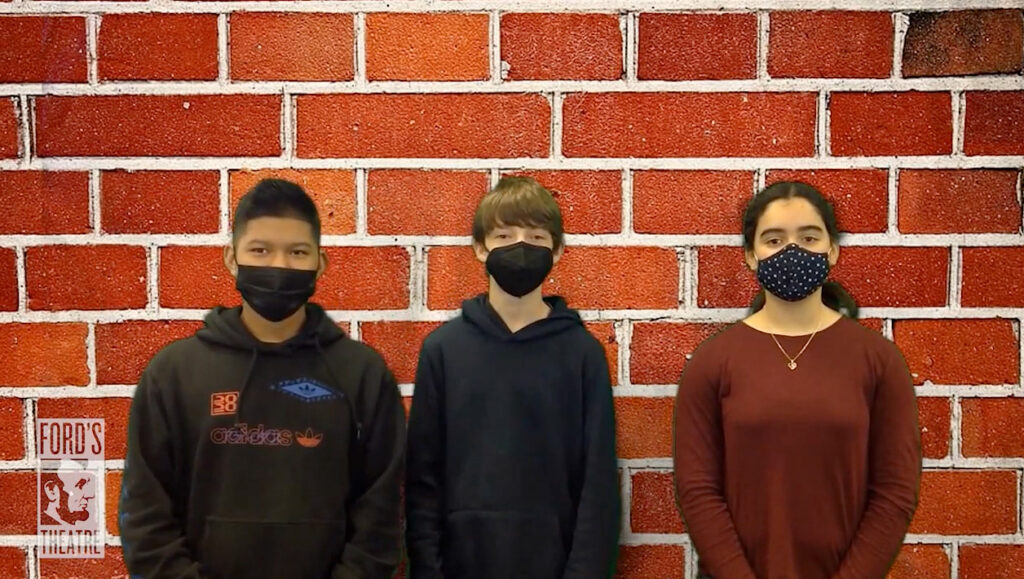 Two young boys and a young girls wearing masks stand in front of a backdrop of a brick wall.