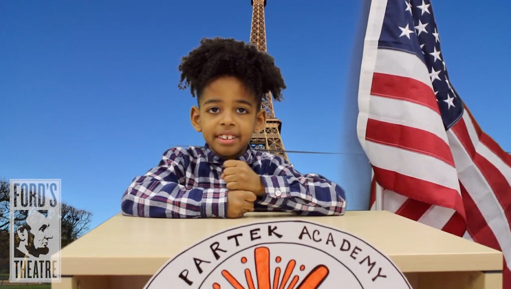 A young boy wearing a plaid shirt sits behind a desk and gives a speech. A sign on the front of the desk reads "Spartek Academy." Behind him is an American flag and a backdrop of the Eiffel tower.