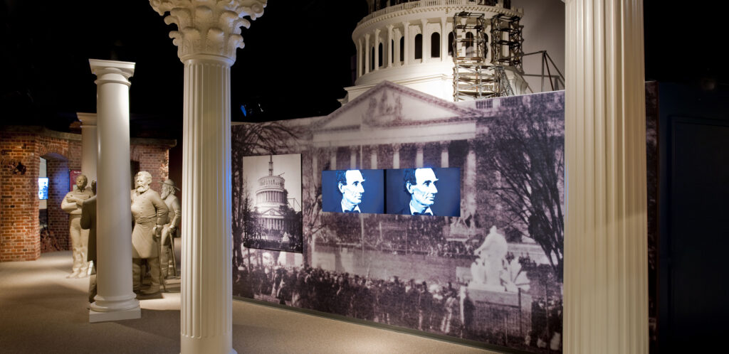 Inside the Ford’s Theatre Museum, two television screens display close-up images of Abraham Lincoln’s face. The screens are embedded in a display that is a large-scale photograph of the partially constructed U.S. Capitol dome taken during the Civil War. In front of the display are three white columns, designed to evoke the Capitol. Behind the display is a model of the partially constructed dome.