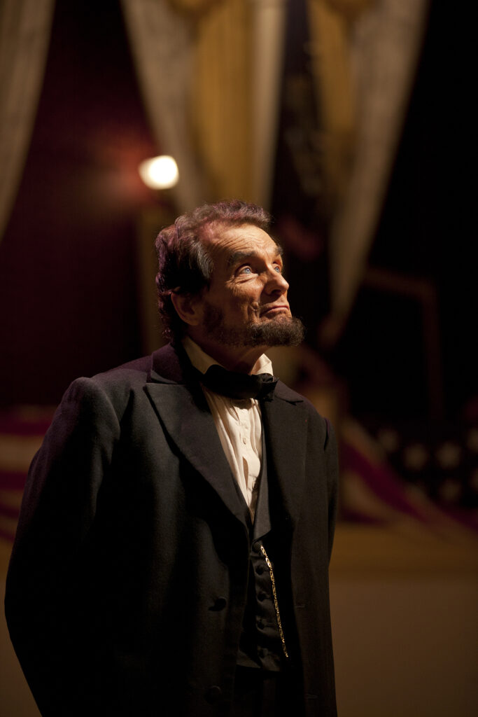 An actor portraying Abraham Lincoln stands and looks above him.