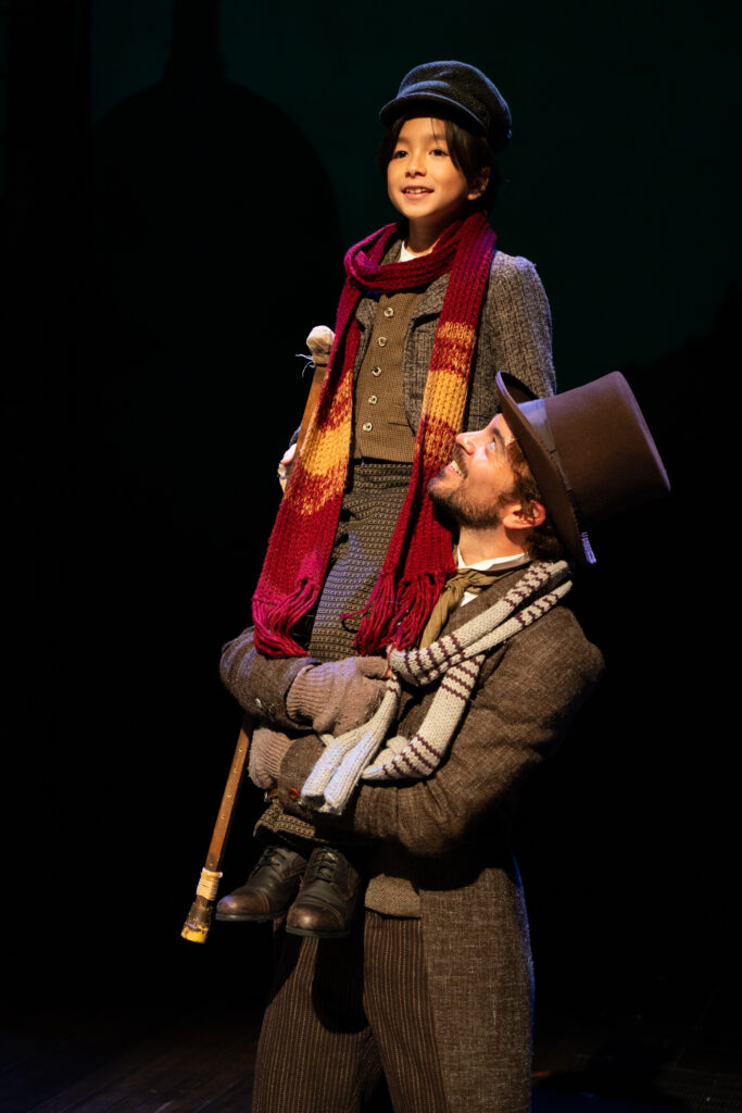 A poor man dressed in worn Victorian-style coat, scarf, fingerless gloves and top hat carries his small child who carries a crutch. Both smile.