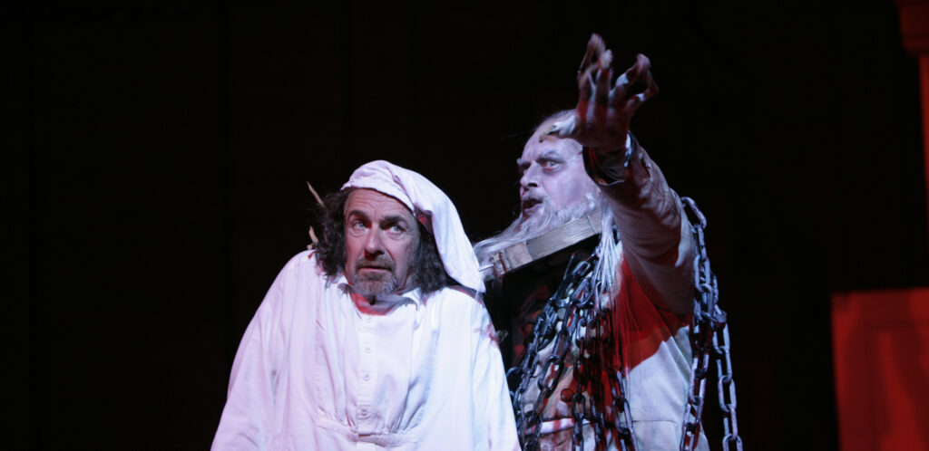 A frightened man in a white nightgown and nightcap stands next to a ghostly figure covered in chains, who gestures towards the camera.