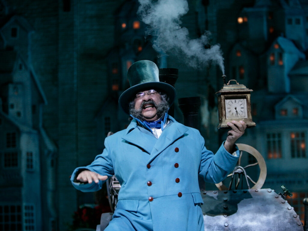 A man dressed in baby-blue, double-breasted Victorian overcoat and black top hat holds a clock and smiles.