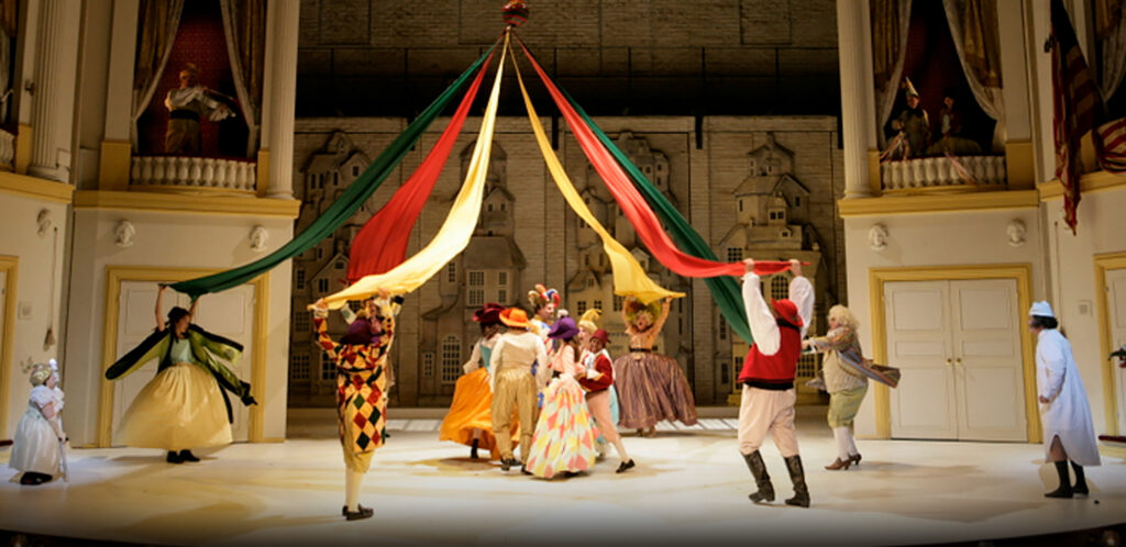 At a party, six people hold colorful fabrics of a May Pole taut as a joyful group of others dance beneath it.