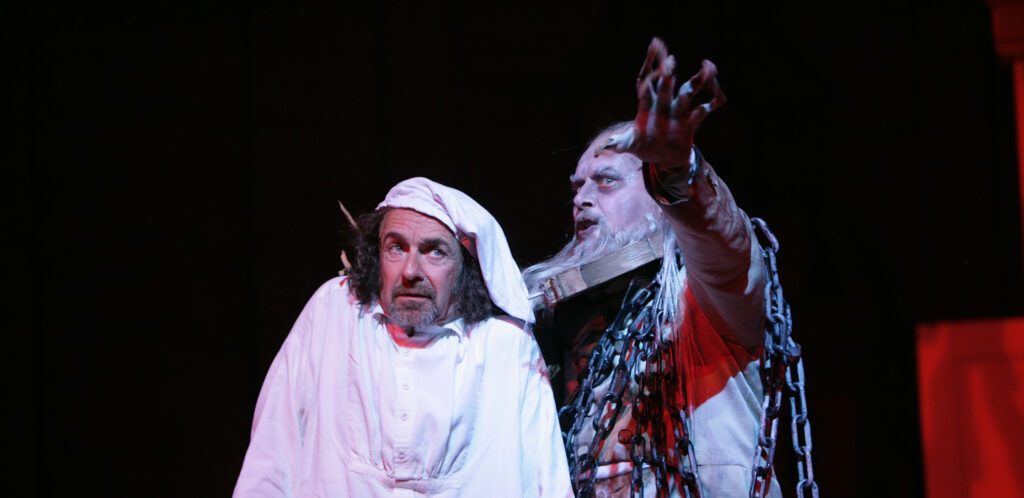 A frightened man in white nightclothes and a white sleeping cap stands next to a ghost wrapped in chains who gestures towards the camera.