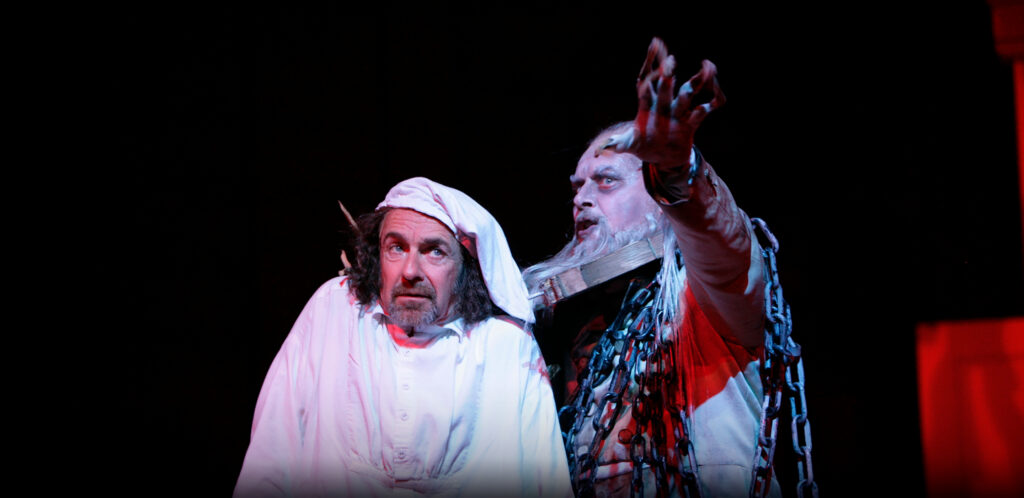 A frightened man in white nightclothes and a white sleeping cap stands next to a ghost wrapped in chains who gestures towards the camera.