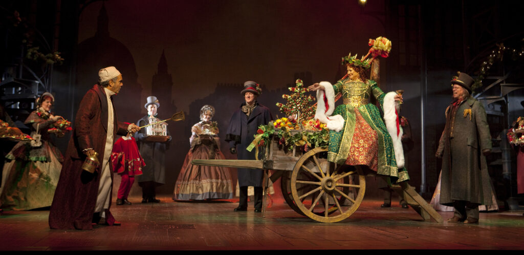 At center is a wooden wagon filled with colorful sparkling fruit and leaves and a small Christmas tree. Upon the wagon sits an older White woman in a brilliantly-colored red, green, and gold gown. The gold designs, piping, and belt of the dress all sparkle, and the sleeves end in floor-length drop sleeves lined with white fur. She wears a green and gold crown with small gold candles set into it. She grins toward a White man standing to her right. The man, Scrooge, wears a white nightcap, a white nightgown, and a dark red silk dressing gown and holds a gold canister in his right hand. The pair are ringed by a group of adults and children, all wearing suits or floor-length gowns and winter cloaks.