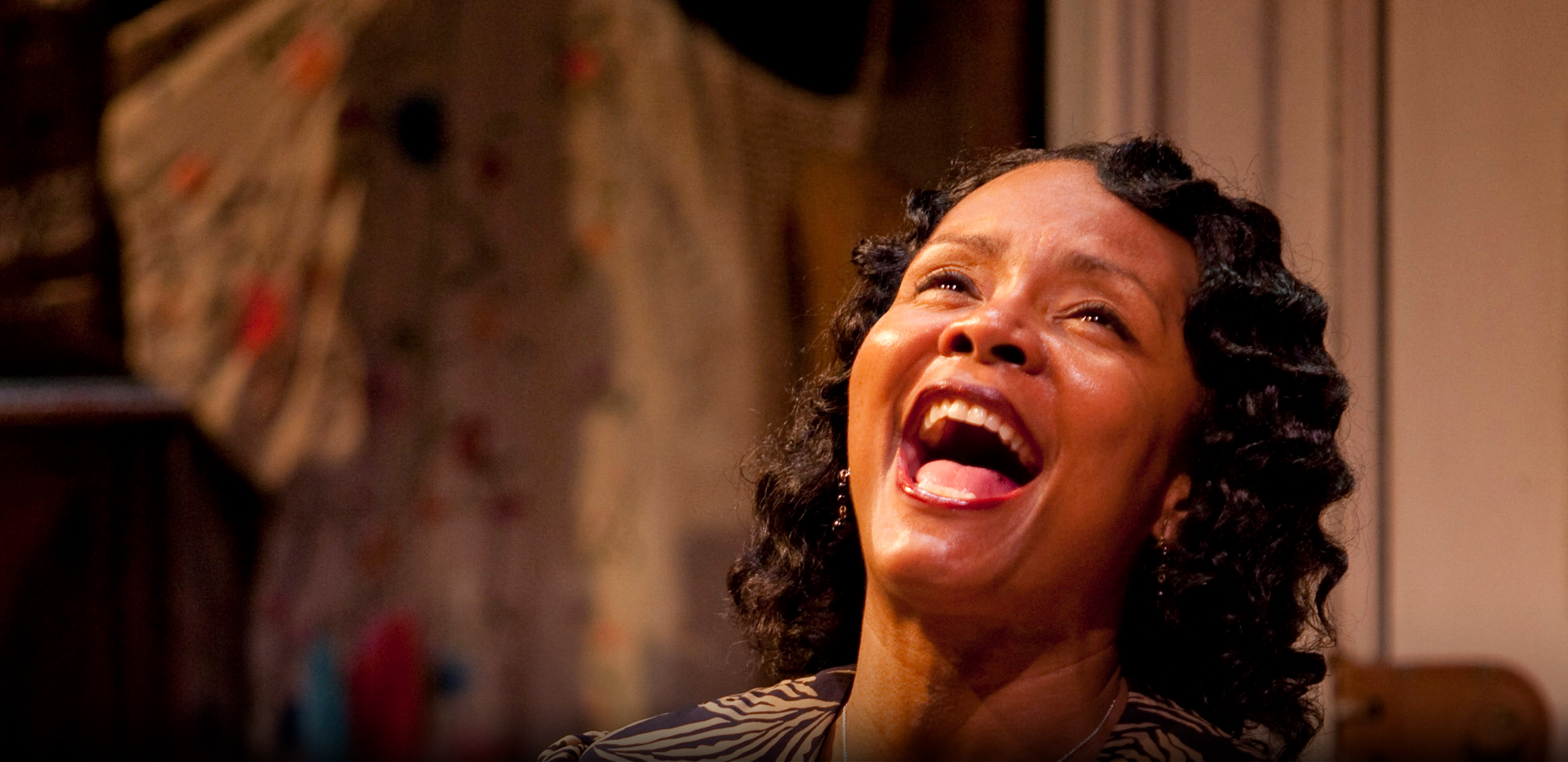 A woman in a blue dress holds a silver bowl of berries in her lap and smiles as she sings.